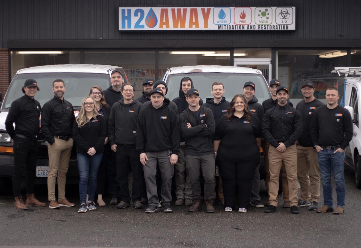 A group of 16 people wearing uniforms stands in front of three white vans and a building with a sign that reads "H2O Away: Water Mitigation and Restoration.